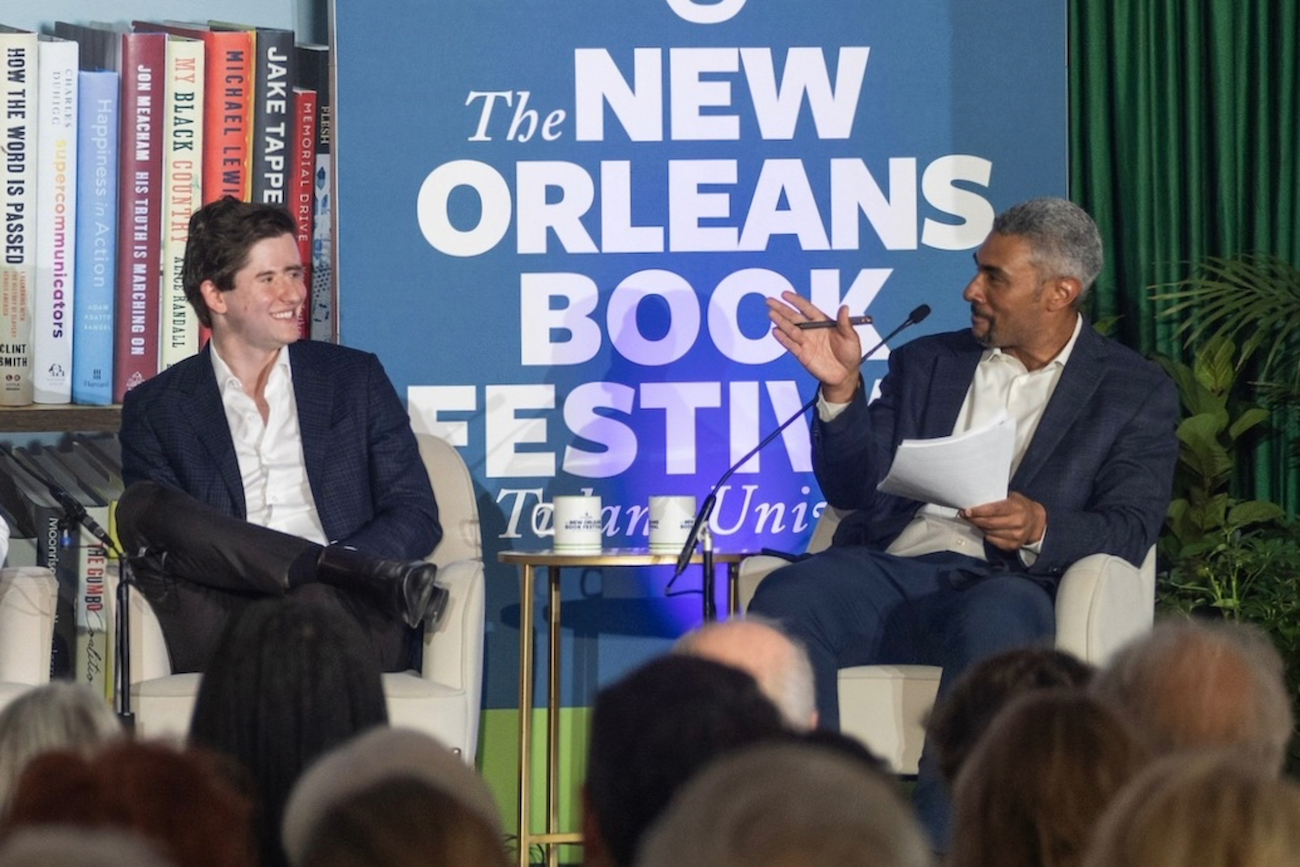 Two men on stage at New Orleans Book Festival. One gestures while speaking, the other smiles.