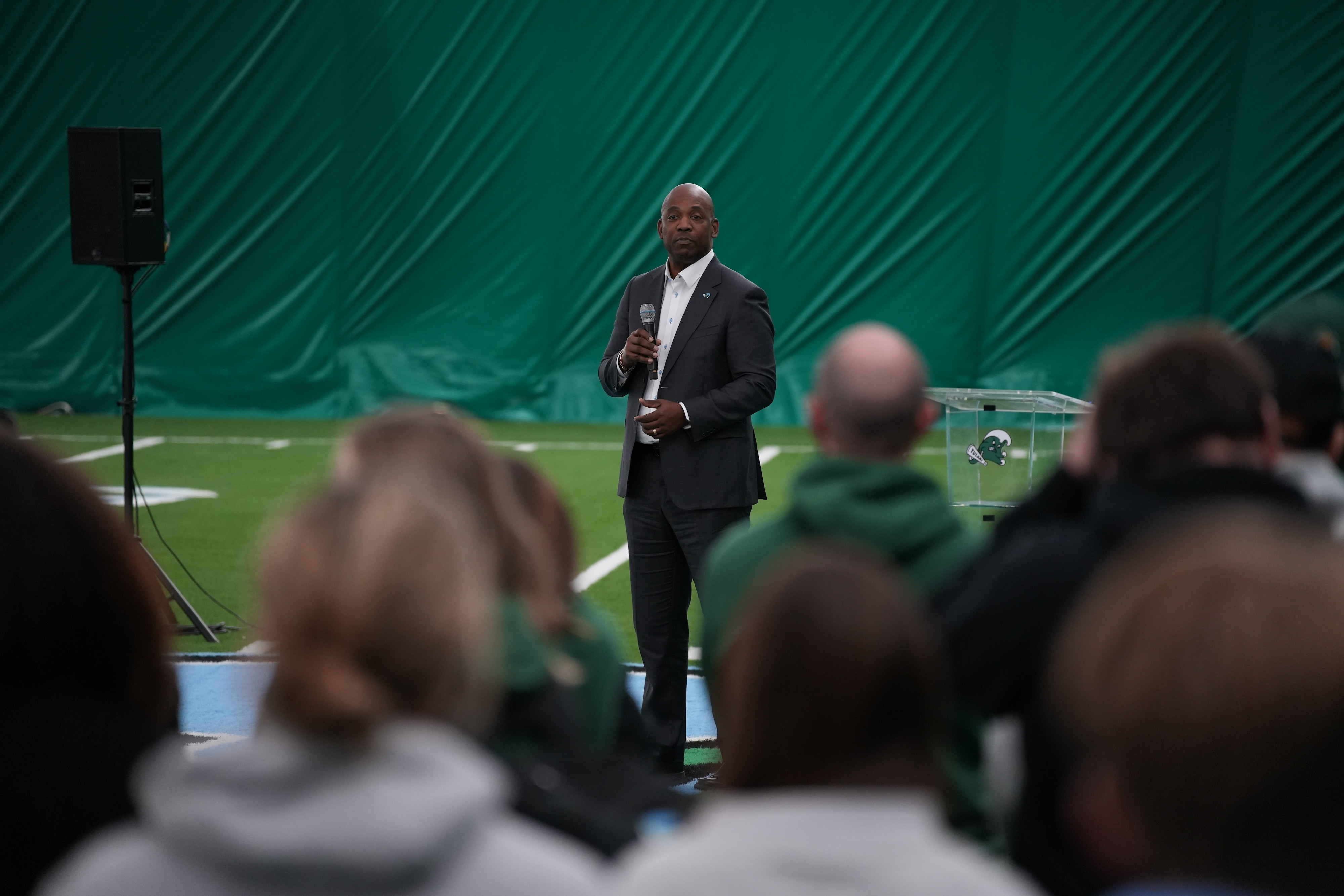 Man in suit speaks into microphone on green turf field to a blurred audience.