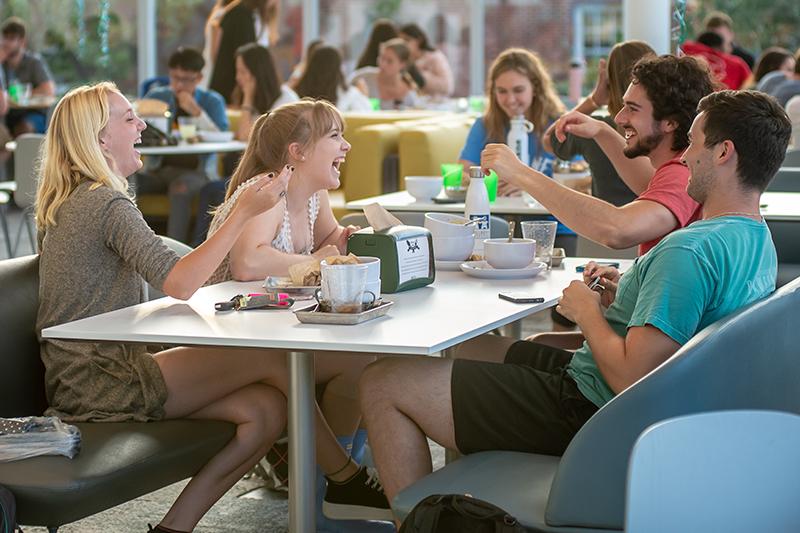 Four friends laugh and socialize at a bright cafeteria table.